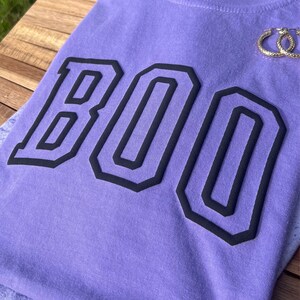 May include: Purple t-shirt with the word "BOO" printed in black outlined letters.