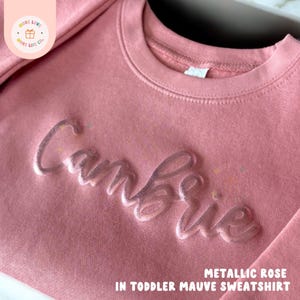 May include: A metallic rose-colored toddler sweatshirt with the name "Cambrie" in a raised, glossy finish. The text "Metallic Rose in Toddler Mauve Sweatshirt" is printed at the bottom. The sweatshirt has a crew neck and long sleeves.