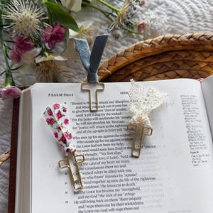 May include: An open book with three decorative bookmarks. One has a blue ribbon, another a floral ribbon, and the third is a white lace design. The text on the page reads "PSALM 94:12".