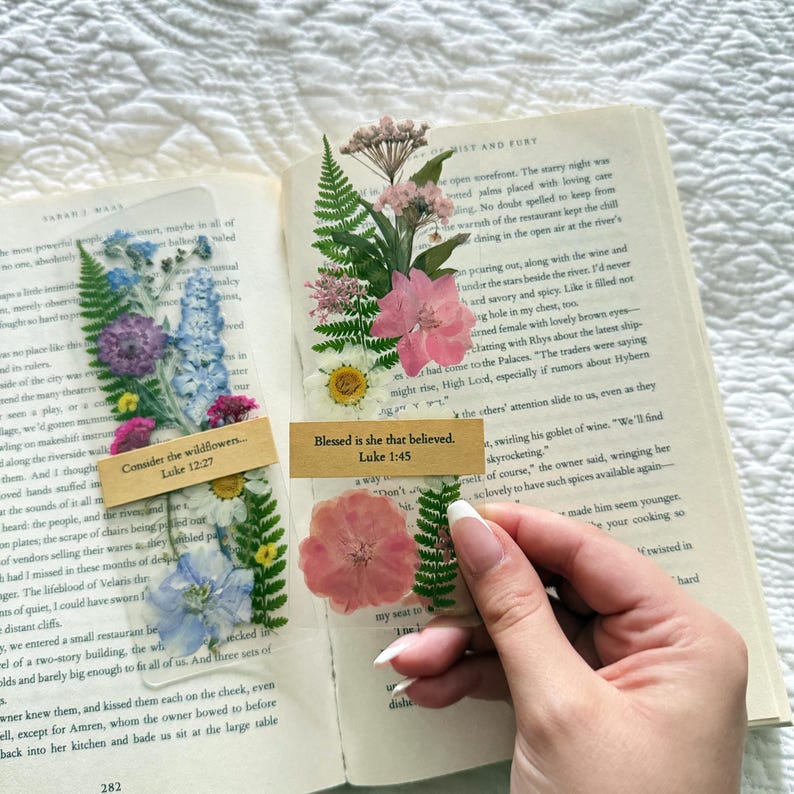 May include: Clear, rectangular bookmarks with pressed flowers and ferns. One bookmark has blue, purple, and white flowers with the text "Consider the wildflowers... Luke 12:27." The other has pink flowers and the text "Blessed is she that believed. Luke 1:45."
