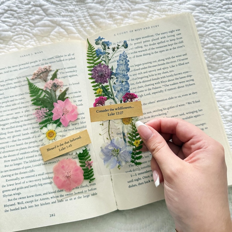 May include: A book with pressed flowers and two clear bookmarks. One bookmark reads "Consider the wildflowers... Luke 12:27" and the other reads "Blessed is she that believed. Luke 1:45". The flowers are pink, purple, blue, and yellow.