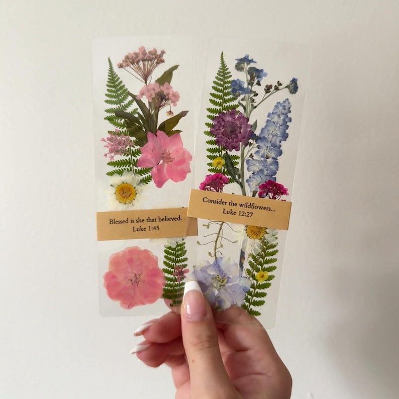May include: Two clear bookmarks with pressed flowers and text. One bookmark has pink and white flowers with the text "Blessed is she that believed. Luke 1:45." The other has purple and blue flowers with the text "Consider the wildflowers... Luke 12:27."