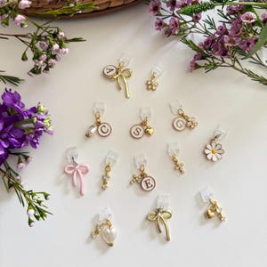 Op de afbeelding: Een verzameling decoratieve bedels in verschillende ontwerpen, waaronder strikken, bloemen, kruisen en initialen. De bedels zijn goudkleurig, roze en wit, met transparante clips. De afbeelding is tegen een witte achtergrond met bloemaccenten.