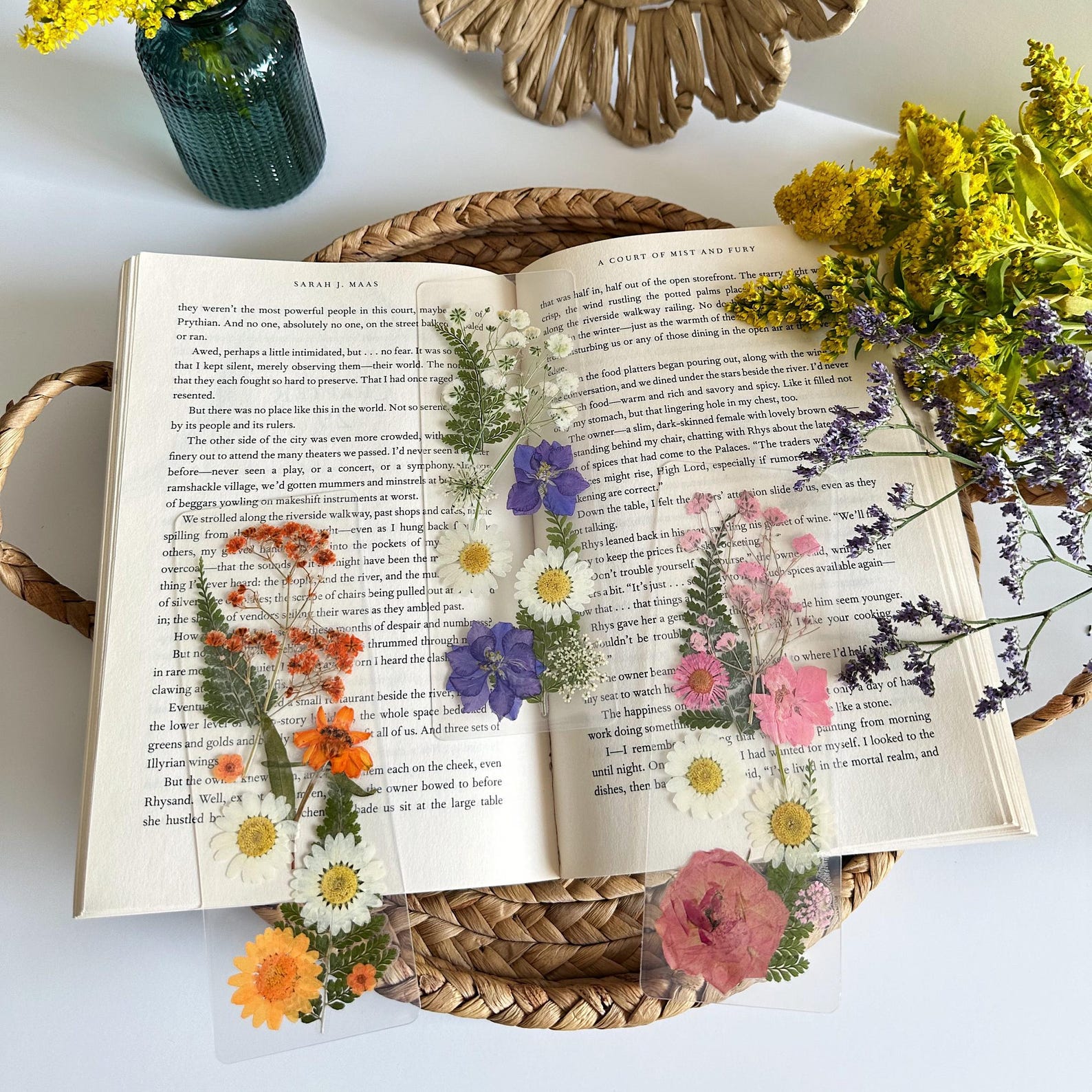 Open book on a tray, with colorful transparent bookmarks with pressed flowers within them. There is a bouquet of purple and yellow flowers on the top right.