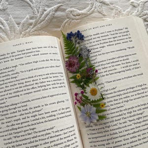 May include: A close-up of an open book with a pressed flower bookmark. The book is titled "Court of Mist and Fury" and the text on the page discusses the differences between the mortal world and the world of Prythian. The bookmark features a variety of pressed flowers, including ferns, daisies, and purple flowers.