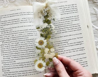 Könnte beinhalten: Transparenter, rechteckiger Lesezeichen mit gepressten weißen und gelben Blumen und grünen Blättern. Oben ist ein weißes Band befestigt. Das Lesezeichen liegt über einem offenen Buch.