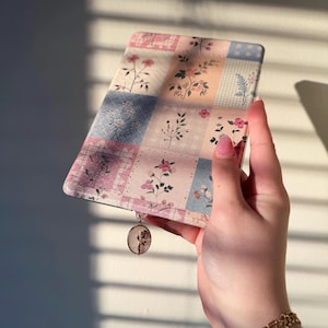 Puede incluir: Una funda de libro con estampado floral en tonos pastel rosa, azul y melocotón. La funda presenta un diseño de patchwork con ilustraciones botánicas. Un pequeño dije con un diseño de rosa cuelga de la parte inferior. La funda se sostiene en una mano.
