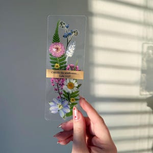 May include: A clear, rectangular bookmark featuring pressed flowers in shades of pink, white, blue, and yellow. A gold-colored band across the middle displays the text "Consider the wildflowers... Luke 12:27".