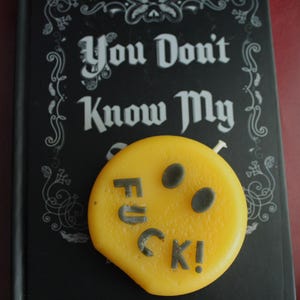 Può includere: Un oggetto giallo e rotondo con la parola "FUCK!" in lettere grigio scuro, posto su un libro nero. La copertina del libro recita "You Don't Know My" in bianco, con un carattere stilizzato. Lo sfondo è rosso scuro.