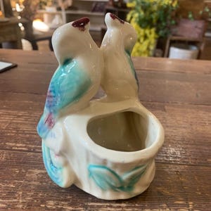May include: A vintage ceramic planter featuring two birds perched together. The birds have white bodies with teal and red accents. The planter has a small opening for plants or flowers and is decorated with teal leaves.