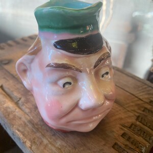 May include: A vintage ceramic pitcher shaped like a man's head. The pitcher features a green hat, black visor, and a pink face with rosy cheeks. The interior is light blue. The pitcher is sitting on a wooden surface.