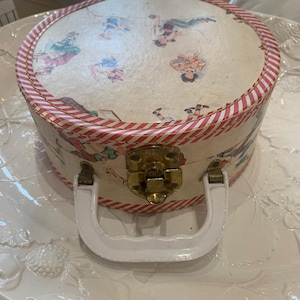 May include: A vintage round hat box with a cream-colored background and illustrations of figures in colorful outfits. The box has a red and white striped border, a white handle, and a brass clasp.