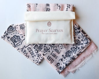 May include: A white cotton pouch with the text "Prayer Scarves" and "This is your secret place."  The pouch contains two pink scarves with black designs and text in multiple languages, including "Jesus" and "Gesù".