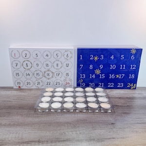 May include: Advent calendar boxes with numbers 1-24, one white and one blue. The white box has a wreath design around the numbers. A clear plastic tray holds 24 small, round, white candles. The boxes are on a wooden surface.