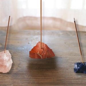 May include: Three natural stone incense holders. One is a light pink rose quartz, another is a red-orange stone, and the third is a dark blue stone. Each holds a thin, brown incense stick. The holders are on a wooden surface.