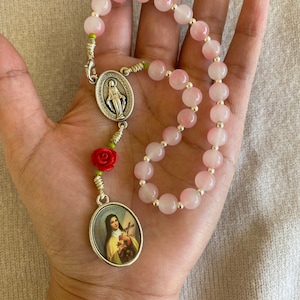 May include: A pink and white rosary with a silver charm featuring an image of the Virgin Mary. The rosary has a red rose charm and a silver clasp.