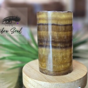 May include: A cylindrical, banded, brown and gold onyx crystal. The crystal is polished and has a smooth, shiny surface. It is sitting on a wooden base.