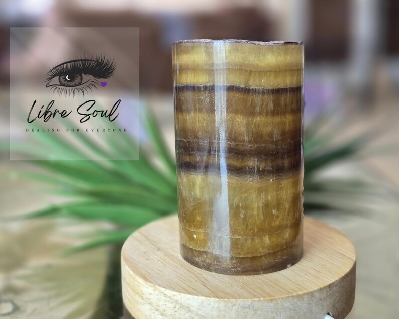 May include: A cylindrical, banded, brown and gold onyx crystal. The crystal is polished and has a smooth, shiny surface. It is sitting on a wooden base.