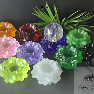 May include: A collection of colorful, flower-shaped candle holders in shades of orange, red, pink, purple, blue, green, and black. A clear crystal ball and a green plant are also present. The candle holders are made of a translucent material.