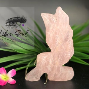 May include: A carved pink rose quartz angel figurine with wings outstretched. The figurine is sitting on a green leafy surface.