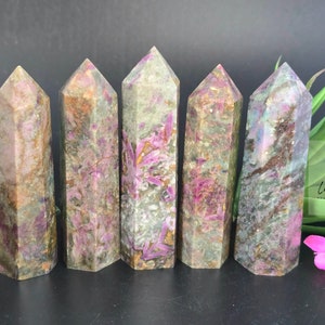 May include: Six pink and green crystal points, also known as rubellite, are arranged in a row on a dark surface. The crystals are all different shapes and sizes, but they all have a similar color pattern. The crystals are surrounded by green foliage and a pink flower.