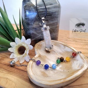 Op de afbeelding: Een zilveren ketting met een heldere kristallen hanger en een zilveren ketting. De hanger is gevuld met kleine witte kristallen en heeft een hartvormig ontwerp aan de bovenkant. De ketting wordt gepresenteerd op een houten hartvormige schaal met een zilveren armband met zeven gekleurde kralen.