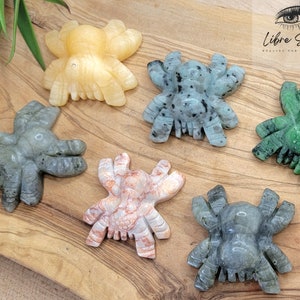 May include: Six carved spider figurines made of various types of stone. The spiders are in different colours, including green, grey, and orange. The spiders are arranged on a wooden surface.