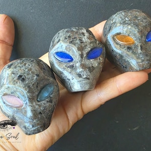 May include: Three alien-shaped carvings made of speckled grey stone. Each carving features smooth, polished surfaces and inlaid almond-shaped eyes in various colours, including blue, pink, and orange. The carvings are held in a palm.