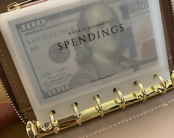 May include: A beige zippered binder featuring a gold-toned six-ring metal mechanism. Inside, a translucent plastic envelope holds a one hundred dollar bill. The envelope is printed with the text WEEKLY EXPENSES SPENDINGS and THERICHBROKEMON.