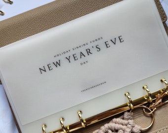 May include: A beige textured binder with gold rings containing a translucent divider labeled New Year's Eve Day and Holiday Sinking Funds. A beige macrame keychain with a tassel is attached to the rings. A white notebook labeled Holiday Savings is visible.