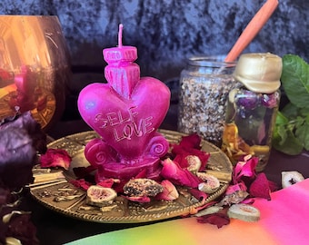 May include: A vibrant pink heart-shaped candle with the words "SELF LOVE" engraved on the front. The candle sits on a decorative gold-colored plate surrounded by dried rose petals. A jar of herbs and a small bottle with a gold seal are in the background.