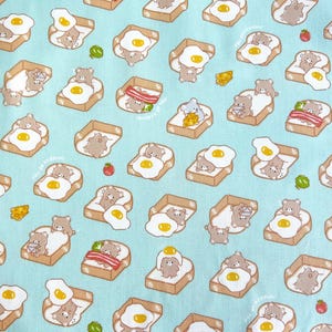 May include: A light blue fabric with a repeating pattern of cartoon bears on toast with fried eggs, bacon, and cheese. The bears are in various poses, some sleeping on the toast. Small red tomatoes and green lettuce are also scattered across the fabric.