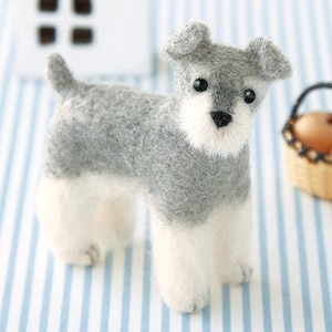 May include: A felt Schnauzer dog figurine in grey and white. The dog has a fluffy texture with detailed features, including small ears, a black nose, and dark eyes. The figurine is standing on a blue and white striped surface.