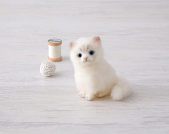 May include: A handmade white cat figurine with blue eyes sits on a light wood surface. A wooden spool of thread and a small ball of twine are next to the cat. The cat has a fluffy appearance.