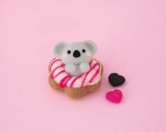 May include: A needle felted gray koala figure sitting inside a brown donut with pink icing and dark pink drizzle. Beside the donut are two small felted hearts, one black and one bright pink, all crafted from soft wool fibers.
