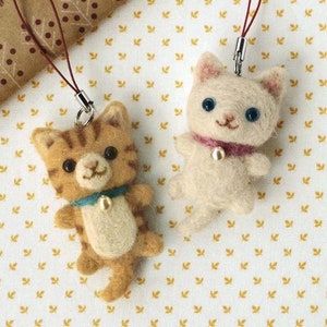 May include: Two handmade felt cat charms. One is tan with brown stripes and a blue collar. The other is white with a pink collar. Both have black eyes and small metal clasps.