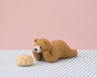 May include: A brown felt bear figurine with a white muzzle and black nose, lying down with its paws near its face. Beside it is a small, round, light-yellow felt object. The background is pink and blue and white striped.
