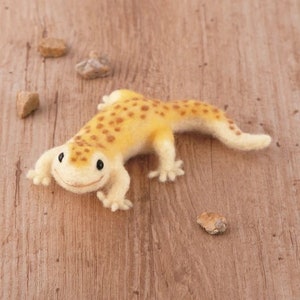 May include: A small, yellow and brown felt gecko with a smiling face. The gecko is lying on a wooden surface.