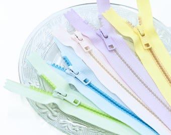 May include: A collection of five nylon coil zippers in pastel shades of mint green, light blue, pale pink, lavender, and soft yellow. Each zipper features a matching plastic slider and pull tab, arranged in a fan shape on a clear glass plate.