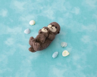 May include: A needle-felted brown sea otter parent holding a small sea otter pup on its chest. The figures have dark brown fur, cream-colored faces, and black eyes. Four small, light-colored seashells are arranged around the otters.