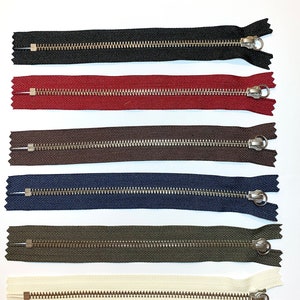 Op de afbeelding: Zes verschillende kleuren metalen ritsen met zilveren tanden. De kleuren zijn zwart, rood, bruin, donkerblauw, olijfgroen en crème.