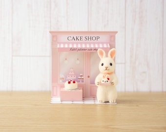 May include: A miniature pink cake shop playset featuring the text CAKE SHOP and Rabbit patissier cake shop. A small flocked white rabbit figurine stands in front, holding a tiny white cake with red cherries on a small tray.