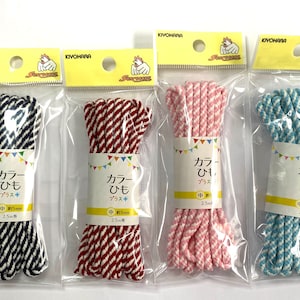 Puede incluir: Cuatro paquetes de cordón de algodón a rayas, cada uno en una combinación de colores diferente: azul marino y blanco, rojo y blanco, rosa y blanco y azul claro y blanco. Cada paquete tiene una etiqueta con el texto "カラーひも プラス+" (japonés para "cordón de colores más") y la longitud del cordón, 2,5 metros.