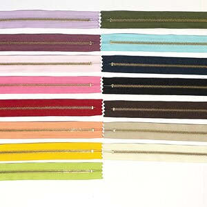 May include: A collection of 10 different colored zippers with gold pulls. The zippers are laid out in two rows of five, with each zipper in a different color. The colors include purple, brown, pink, red, orange, yellow, green, blue, black, and beige.