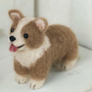 Puede incluir: Un juguete de corgi de fieltro de aguja marrón y blanco. El corgi tiene un pecho blanco y una lengua rosa que sobresale.