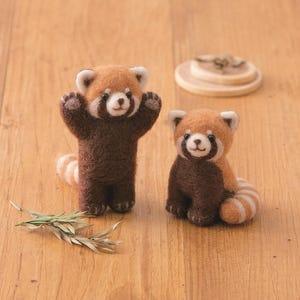 May include: Two charming red panda plushies crafted from felt. The larger panda is standing with its arms raised, while the smaller panda is sitting with its tail curled around its legs. Both pandas have brown fur with white markings on their faces and tails.