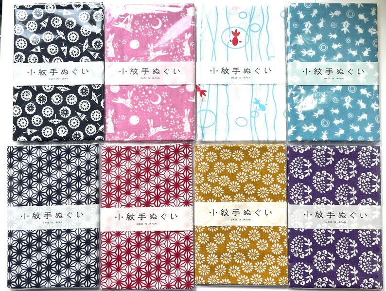 May include: Eight rectangular fabric cloths with diverse patterns including black umbrellas, pink rabbits, blue fish, and floral motifs. Each cloth is wrapped in a white paper band featuring the Japanese text 小紋手ぬぐい and MADE IN JAPAN.