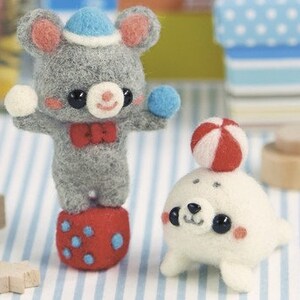 Könnte beinhalten: Eine graue Filzmaus mit einem blauen Hut und einer roten Schleife balanciert auf einem roten und blauen Filzwürfel. Ein weißes Filzrobbe mit schwarzen Augen und einem roten und weißen Ball auf dem Kopf sitzt auf einer gestreiften Oberfläche.