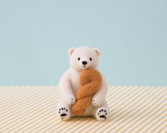 May include: A handmade white polar bear figurine with black eyes and paws, holding a brown pretzel-shaped object. The bear is sitting on a yellow and white striped surface, with a light blue background.