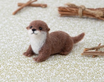 May include: A small, brown and white needle felted otter figurine. The otter is lying on its belly with its head turned to the side and its eyes looking at the camera.